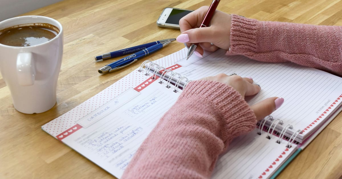 A hand writing figures in a ruled notebook beside a mug of coffee