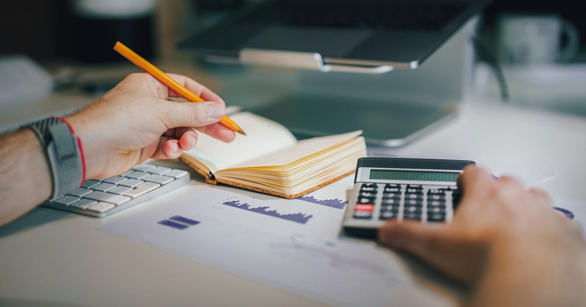 A hand writing in a notebook next to a desk calculator and a pencil