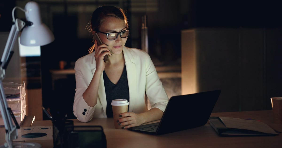 A person working late at a laptop with a phone held to one ear and a coffee cup alongside