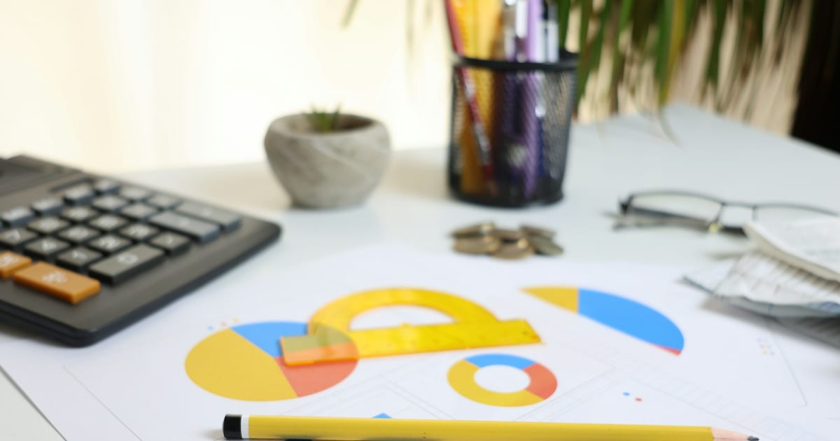 A desk calculator resting on printed charts beside a pencil and a pair of glasses