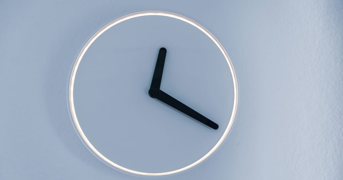 A plain wall clock with black hands against a pale grey wall